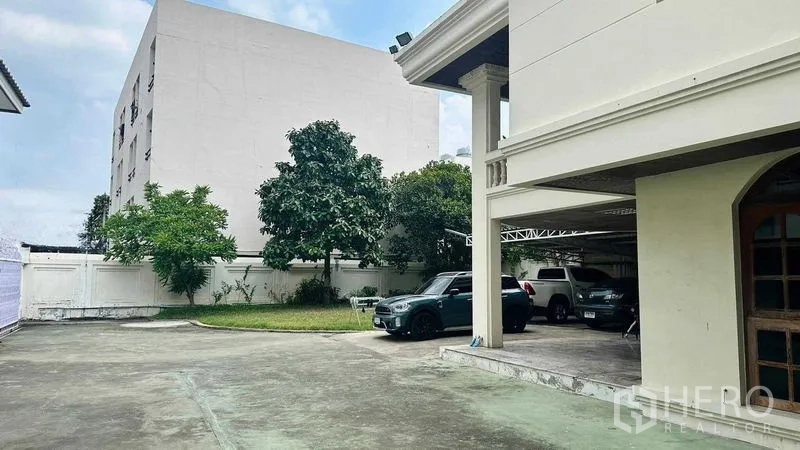 Detached House for sale in Huai Khwang, Bangkok - Spacious concrete driveway with lawn and mature trees beside a detached house in Huai Khwang, Bangkok, with covered parking.