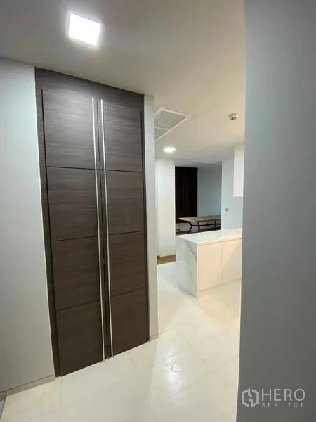 Condominium for rent or sale in Khlong Toei, Bangkok - Entry corridor with tall dark wood cabinet leading to a bright white kitchen island.