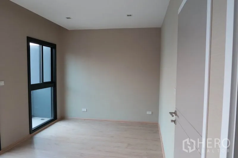 Townhouse for rent in Suan Luang, Bangkok - Bedroom with window and door to a small balcony, light wood floors, and soft lighting.