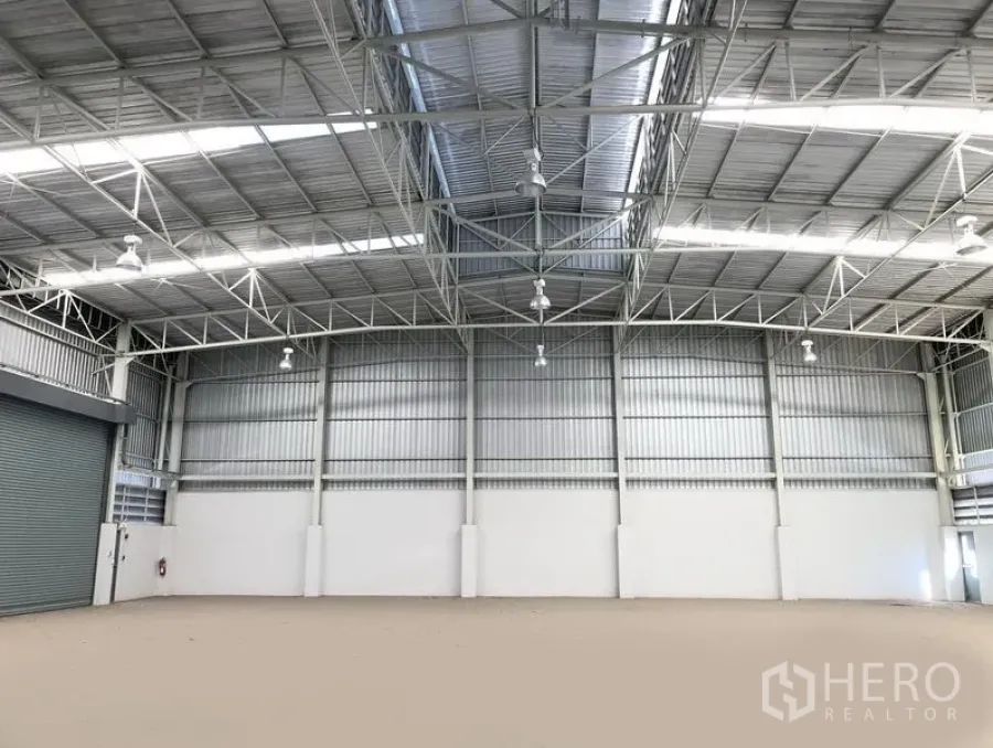 Factory for rent or sale in Bang Sao Thong, Samut Prakan - High-clearance factory warehouse interior with steel truss roof, skylights, bright high-bay lights and a roller shutter door.