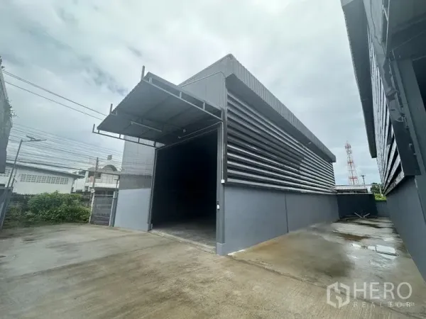 货栈 出租在Prawet, Bangkok - 现代灰色仓库外立面，带遮雨装卸区与宽阔混凝土车道，曼谷Prawet。