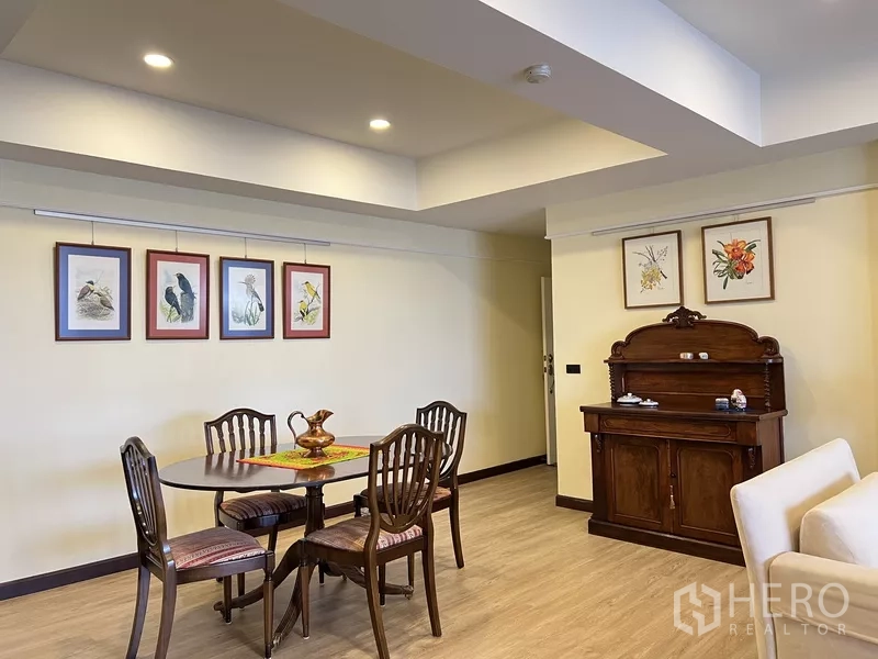Condominium for rent in Khlong Toei, Bangkok - Dining area with round wooden table, four chairs and framed artwork under ceiling spotlights.