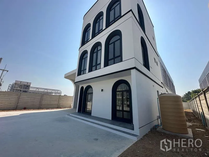 工厂 出售在Lam Luk Ka, Pathum Thani - 建筑侧立面，拱形窗、硬化场地与围墙旁的水塔。