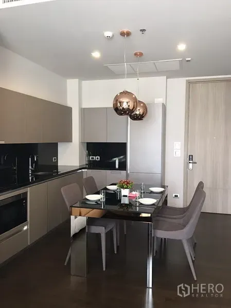 Condominium for rent in Watthana, Bangkok - Open-plan kitchen and dining area with black glass-top table set for four and copper pendant lights.