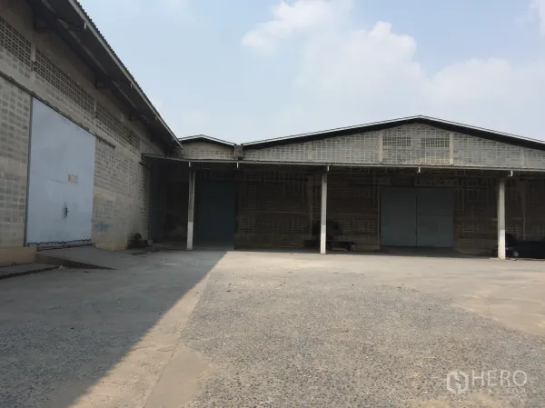 Warehouse for rent in Mueang Pathum Thani, Pathum Thani - Single-storey warehouse exterior with large sliding metal doors, covered loading bay and wide concrete yard in Mueang Pathum Thani.