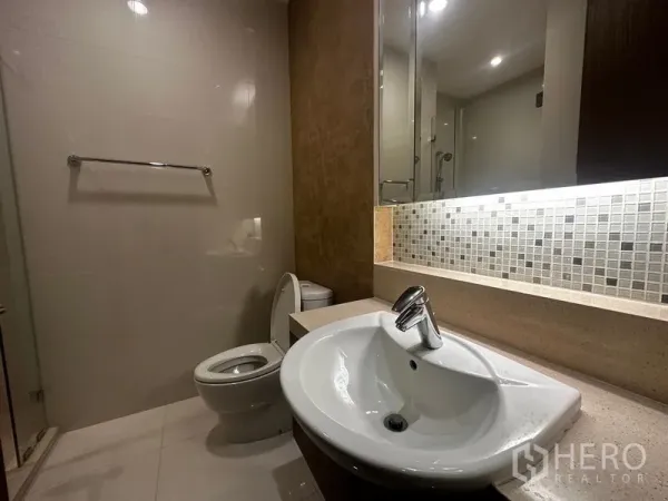 Condominium for rent in Khlong Toei, Bangkok - Modern bathroom with illuminated mirror, sink, toilet and glass shower zone.