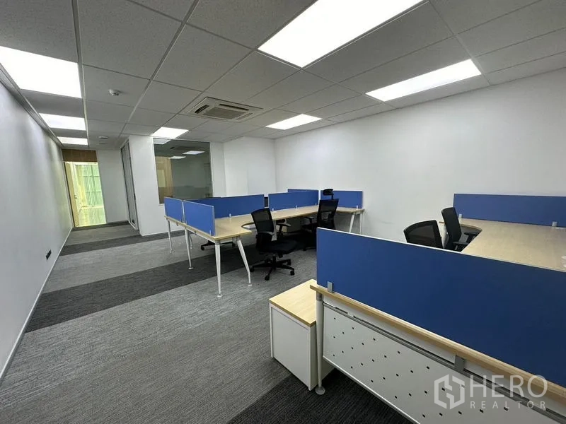 Office Space for rent - Workstation area from another angle showing multiple desks, blue dividers, and corridor to glass door.