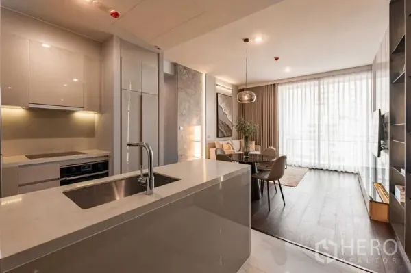 Condominium for rent in Watthana, Bangkok - Open-plan kitchen with white island and sink leading to a bright living and dining area with full-height curtains.