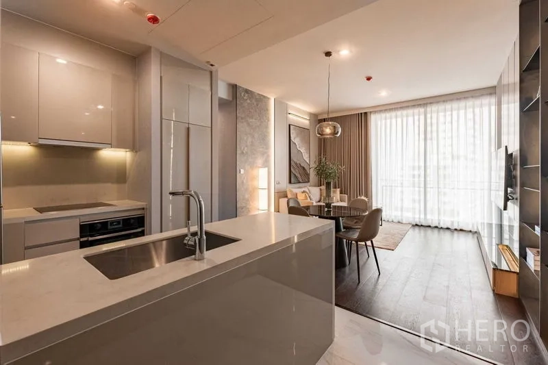 Condominium for rent in Watthana, Bangkok - Open-plan kitchen with white island and sink leading to a bright living and dining area with full-height curtains.