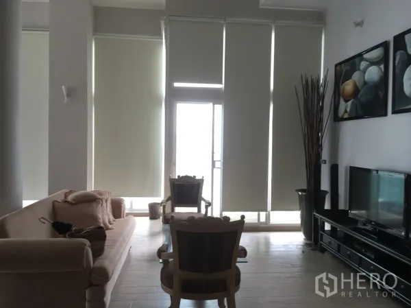 Condominium for rent or sale in Watthana, Bangkok - Spacious high‑ceiling living room with floor‑to‑ceiling windows, roller blinds, sofa set and TV console opening to a balcony.