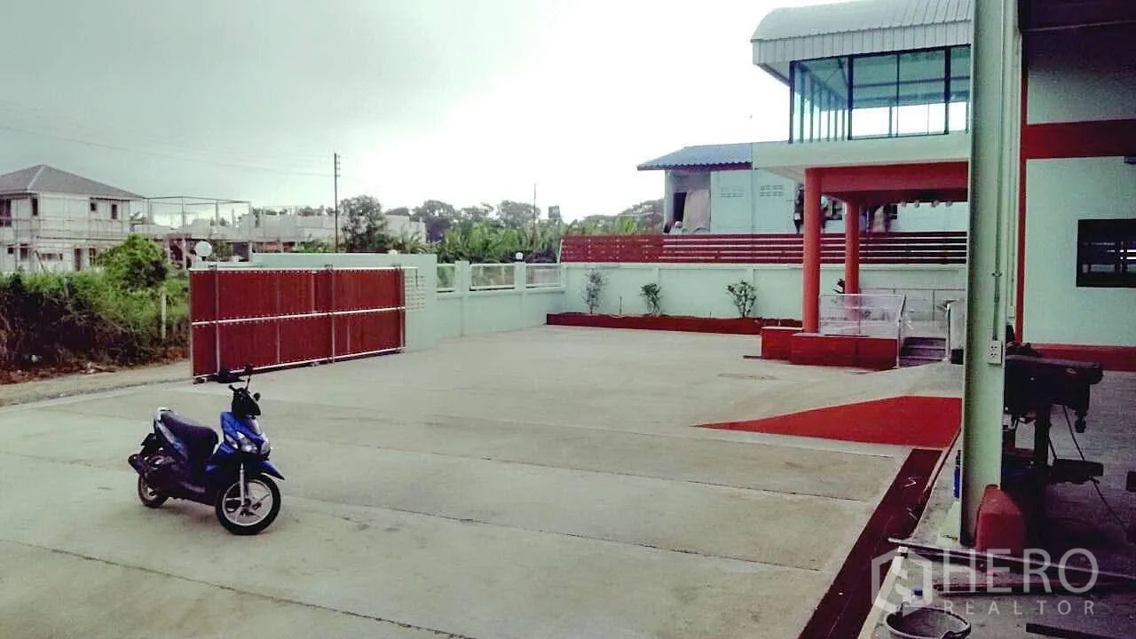 Factory for sale in Pathum Thani - Wide concrete yard with sliding gate and parking area in front of the factory.