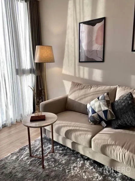 Condominium for rent or sale in Phaya Thai, Bangkok - Cozy seating corner with beige sofa, side table and standing lamp beside sheer curtains.