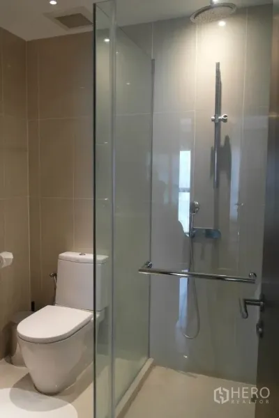 Condominium for rent or sale in Sathon, Bangkok - Bathroom with glass‑enclosed rain shower and toilet in neutral tiles.