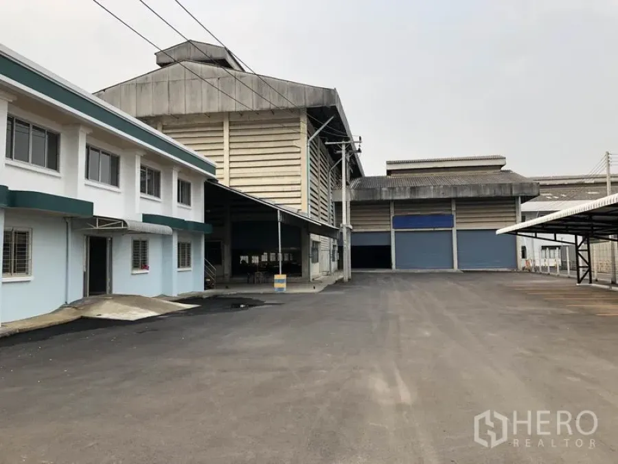 Factory for rent in Lam Luk Ka, Pathum Thani - Wide asphalt yard between office block and warehouses with covered loading and roller doors.