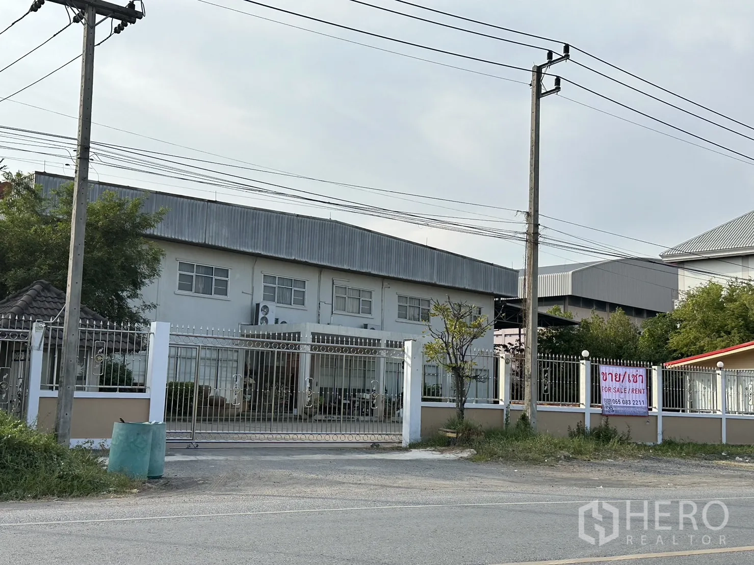Factory for rent or sale in Lam Luk Ka, Pathum Thani - Street view of a gated factory complex with a two‑storey office building and power lines in Lam Luk Ka.