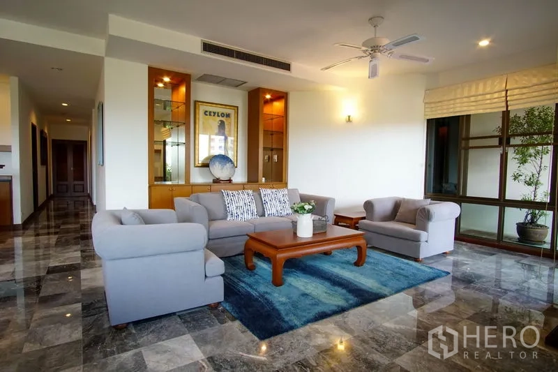Apartment for rent in Watthana, Bangkok - Bright living room with grey sofa set, blue rug, built-in display cabinets, and large windows.