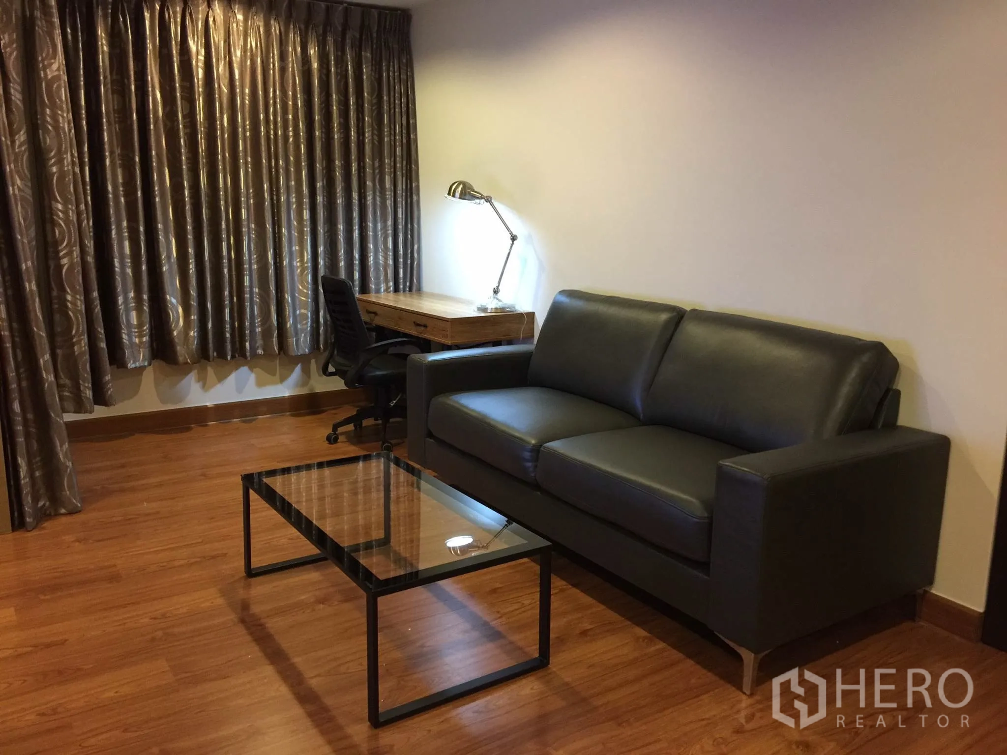 公寓 出租在Bang Rak, Bangkok - 客厅配黑色皮沙发、玻璃茶几及窗边书桌，厚窗帘遮光