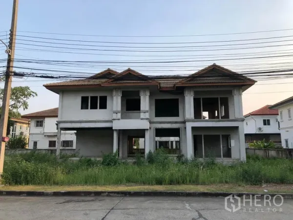 Detached House for sale in Lam Luk Ka, Pathum Thani - Front view of an unfinished two-storey detached house with red-tiled roof and overgrown lawn in Lam Luk Ka, Pathum Thani.