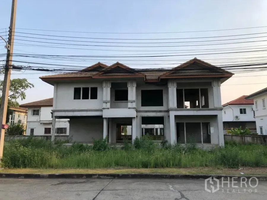 Detached House for sale in Lam Luk Ka, Pathum Thani - Front view of an unfinished two-storey detached house with red-tiled roof and overgrown lawn in Lam Luk Ka, Pathum Thani.