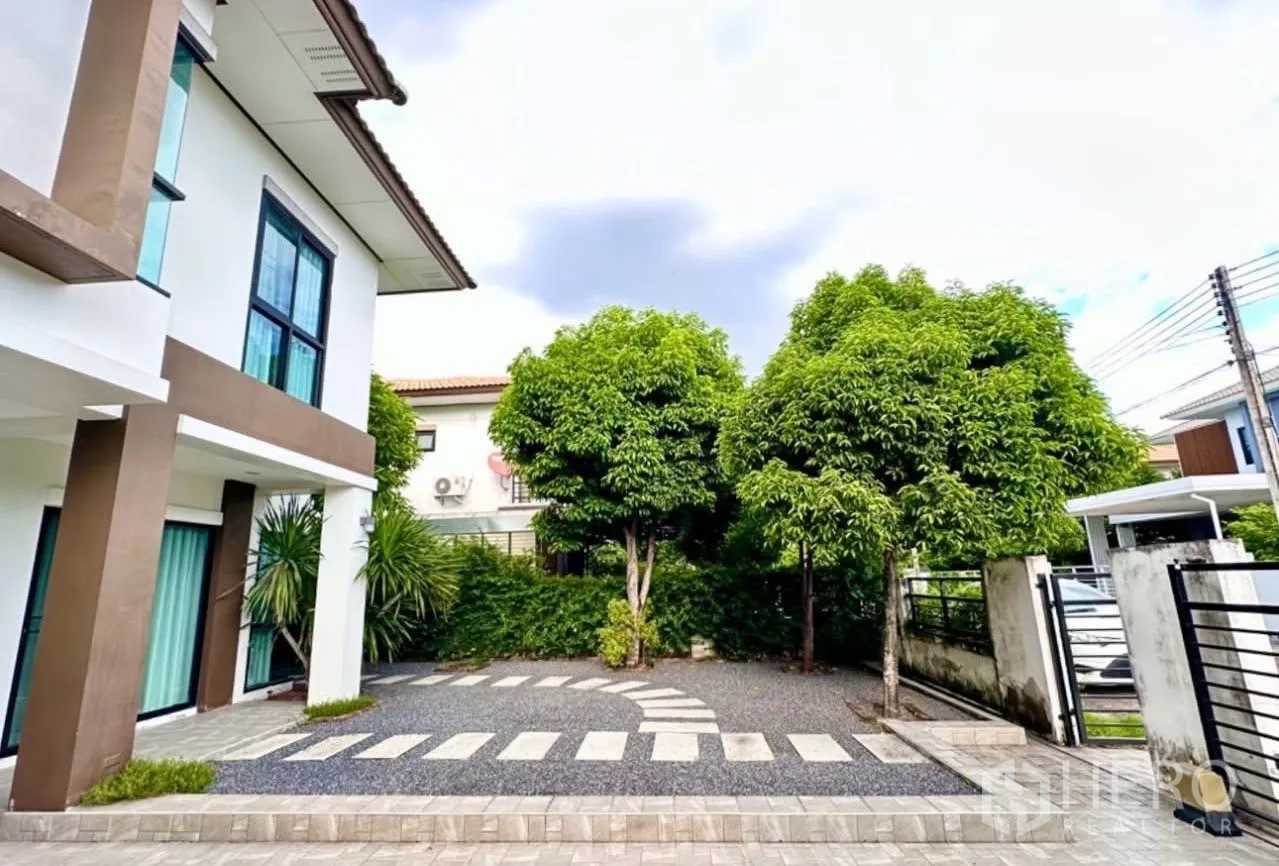Detached House for sale in Lam Luk Ka, Pathum Thani - Front yard showing paved driveway, stepping stones, shade trees and gate entrance.