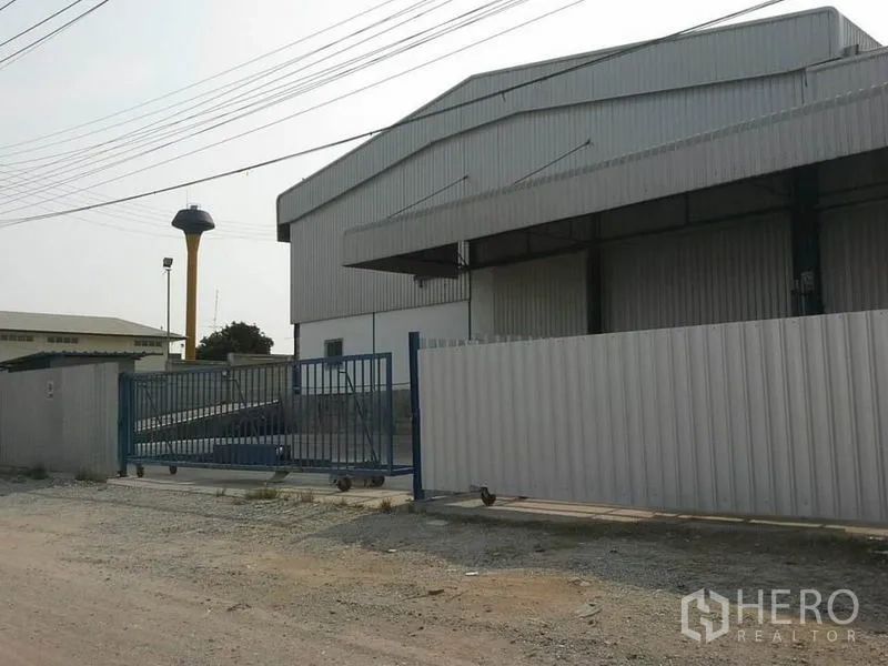 Warehouse for rent in Chon Buri - Warehouse exterior with perimeter fence and sliding gate, wide road access and nearby water tower.