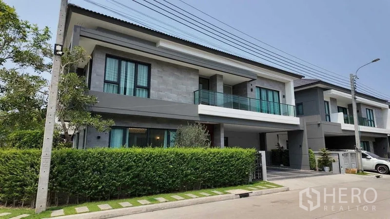 บ้าน ให้เช่าหรือขายในBang Na, Bangkok - ด้านหน้าบ้านเดี่ยวมองจากถนน พร้อมรั้วพุ่มไม้และระเบียงกระจก
