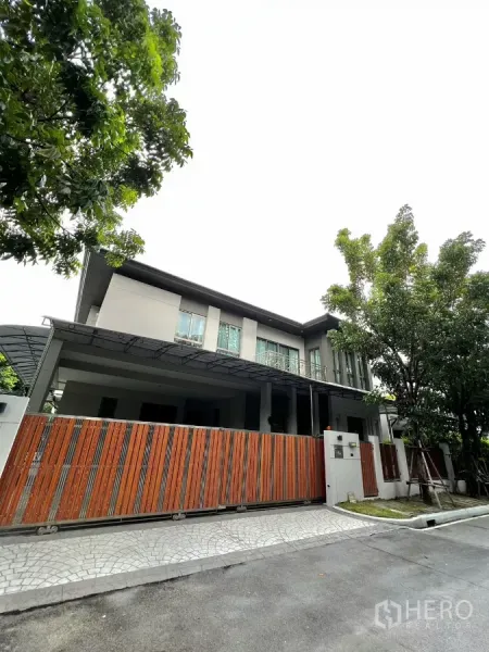 Detached House for rent or sale in Prawet, Bangkok - Modern two-storey detached house with tall trees and a wooden sliding gate on a quiet lane.