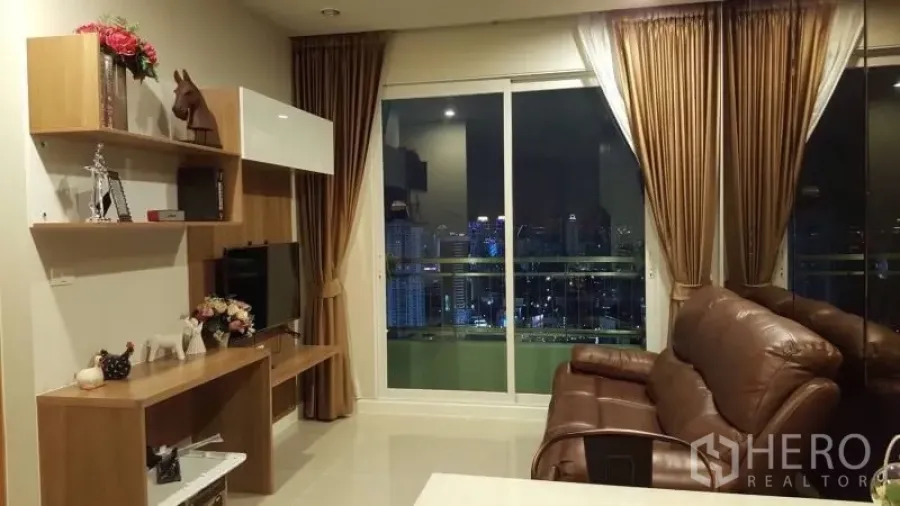 คอนโด ให้เช่าหรือขายในRatchathewi, Bangkok - ห้องนั่งเล่นพร้อมโซฟาหนัง ชั้นวางทีวีบิวท์อิน และประตูกระจกบานเลื่อนออกสู่ระเบียงชมวิวสกายไลน์กรุงเทพฯ ยามค่ำคืน