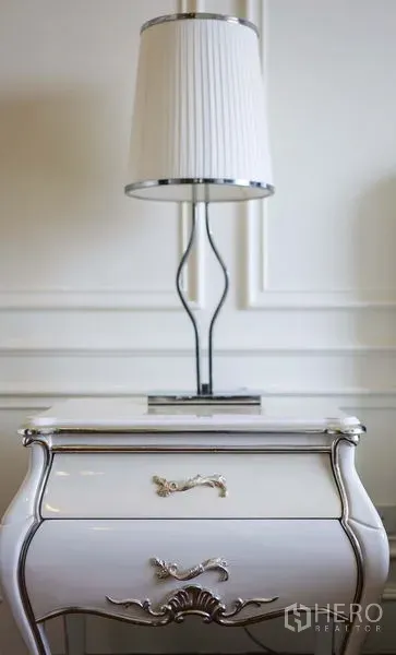 Condominium for rent in Khlong Toei, Bangkok - Close-up of white bedside table with chrome lamp and decorative wall paneling.