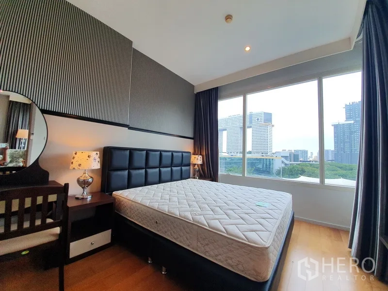 Condominium for rent in Chatuchak, Bangkok - Bedroom with large window, upholstered headboard bed, desk and night lamps, city skyline beyond.