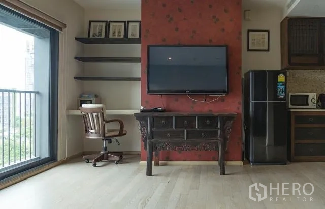 Condominium for rent in Khlong Toei, Bangkok - Living area with wall-mounted TV, study desk and shelves, opening to a balcony city view.