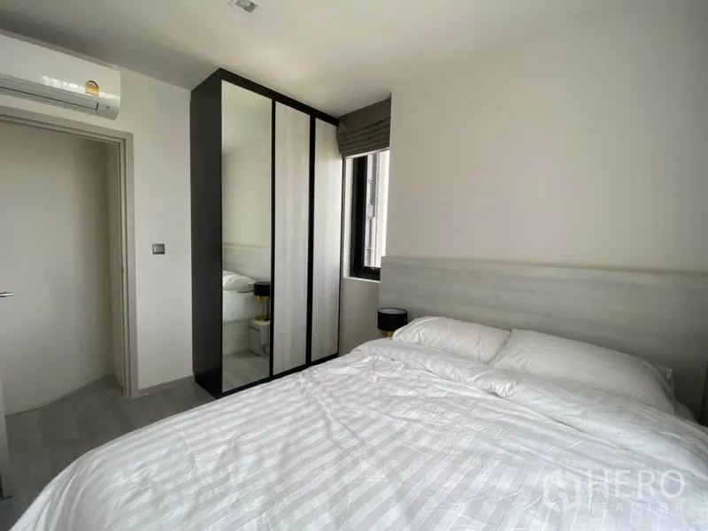 Condominium for rent in Phra Khanong, Bangkok - Bedroom angle showing mirrored wardrobe, air conditioning unit, side window and bedside table.