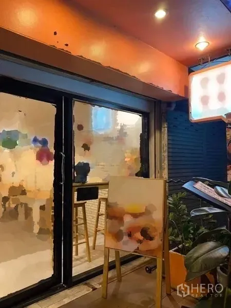 店屋 出租在Phaya Thai, Bangkok - 夜间店面视角，橙色雨棚下的玻璃入口、招牌与绿植。