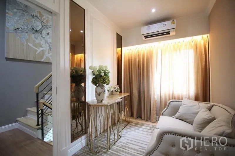 Detached House for rent or sale in Lat Phrao, Bangkok - Upstairs landing lounge with sofa, decorative console table and staircase, warmly lit by curtains.