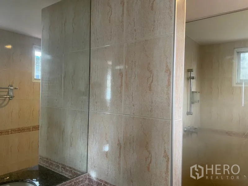 บ้าน ให้เช่าในHuai Khwang, Bangkok - ห้องน้ำกรุกระเบื้องสีเบจ ฉากกั้นกระจก เคาน์เตอร์อ่างล้างหน้า และหน้าต่างระบายอากาศ.