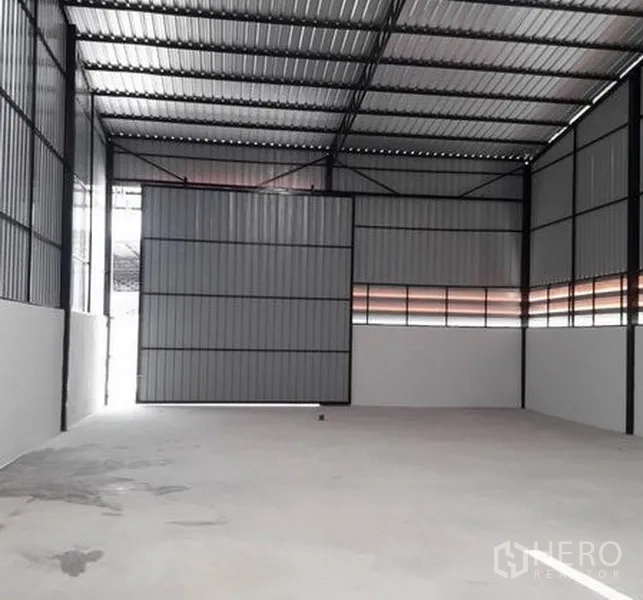 Warehouse for rent in Lam Luk Ka, Pathum Thani - Spacious warehouse with a tall metal sliding door partly open, corrugated steel walls and concrete floor.
