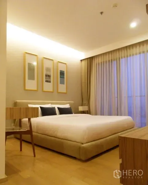 คอนโด ให้เช่าในPhaya Thai, Bangkok - ห้องนอนโทนอุ่น เตียงบุผ้า ภาพแขวน และผ้าม่านสูงจรดเพดานเปิดสู่ระเบียง