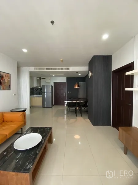 公寓 出租在Watthana, Bangkok - 开放式客餐厅视角，橙色沙发与茶几，后方可见厨房与冰箱。