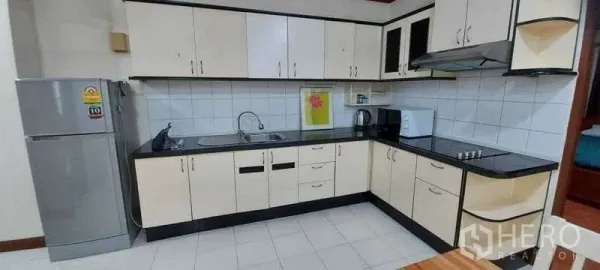 Condominium for rent in Khlong Toei, Bangkok - L‑shaped kitchen featuring white cabinetry, black countertops, stovetop, hood, fridge and microwave.
