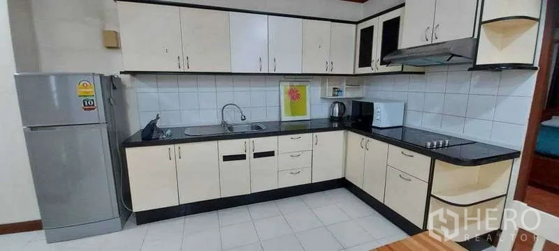 Condominium for rent in Khlong Toei, Bangkok - L‑shaped kitchen featuring white cabinetry, black countertops, stovetop, hood, fridge and microwave.