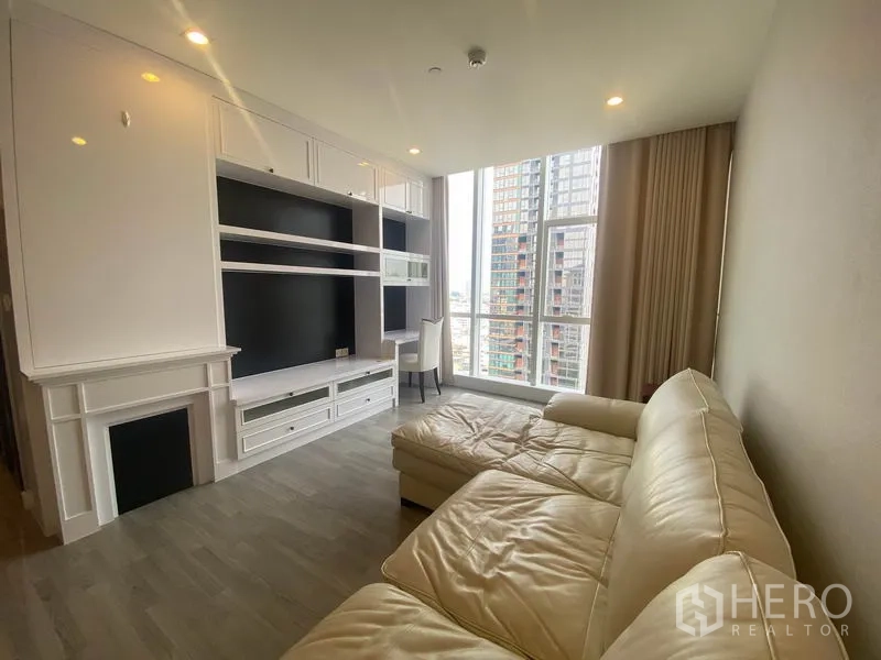 Condominium for rent in Bang Rak, Bangkok - Bright living room with white built-in media wall, L-shaped sofa and floor-to-ceiling windows with Bangkok skyline view.