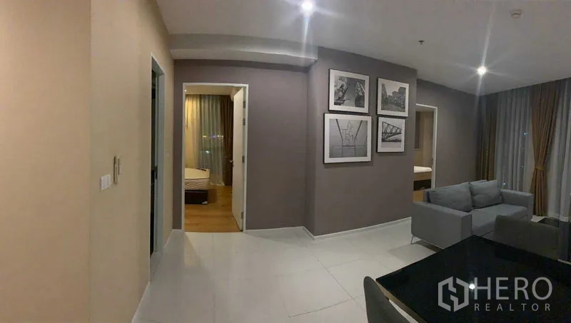 公寓 出租在Watthana, Bangkok - 走廊通往两间卧室，墙面装饰画，瓷砖地面，旁为带大窗的起居区。
