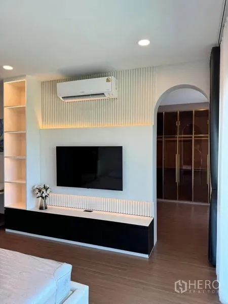 Detached House for rent or sale in Bang Phli, Samut Prakan - Bedroom media wall with TV, LED-lit slatted panel, air conditioner, and an arched doorway to the walk-in closet.