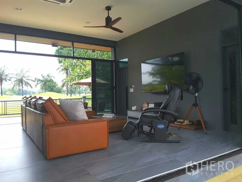Detached House for rent or sale in Lam Luk Ka, Pathum Thani - Open living room with leather sofa, ceiling fan and large TV wall, sliding doors to golf fairway view.