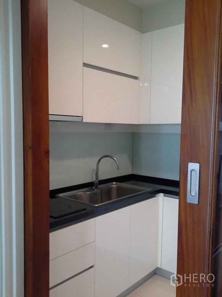 Condominium for rent or sale in Khlong Toei, Bangkok - Compact L-shaped kitchen with stainless sink and a sliding wooden door.