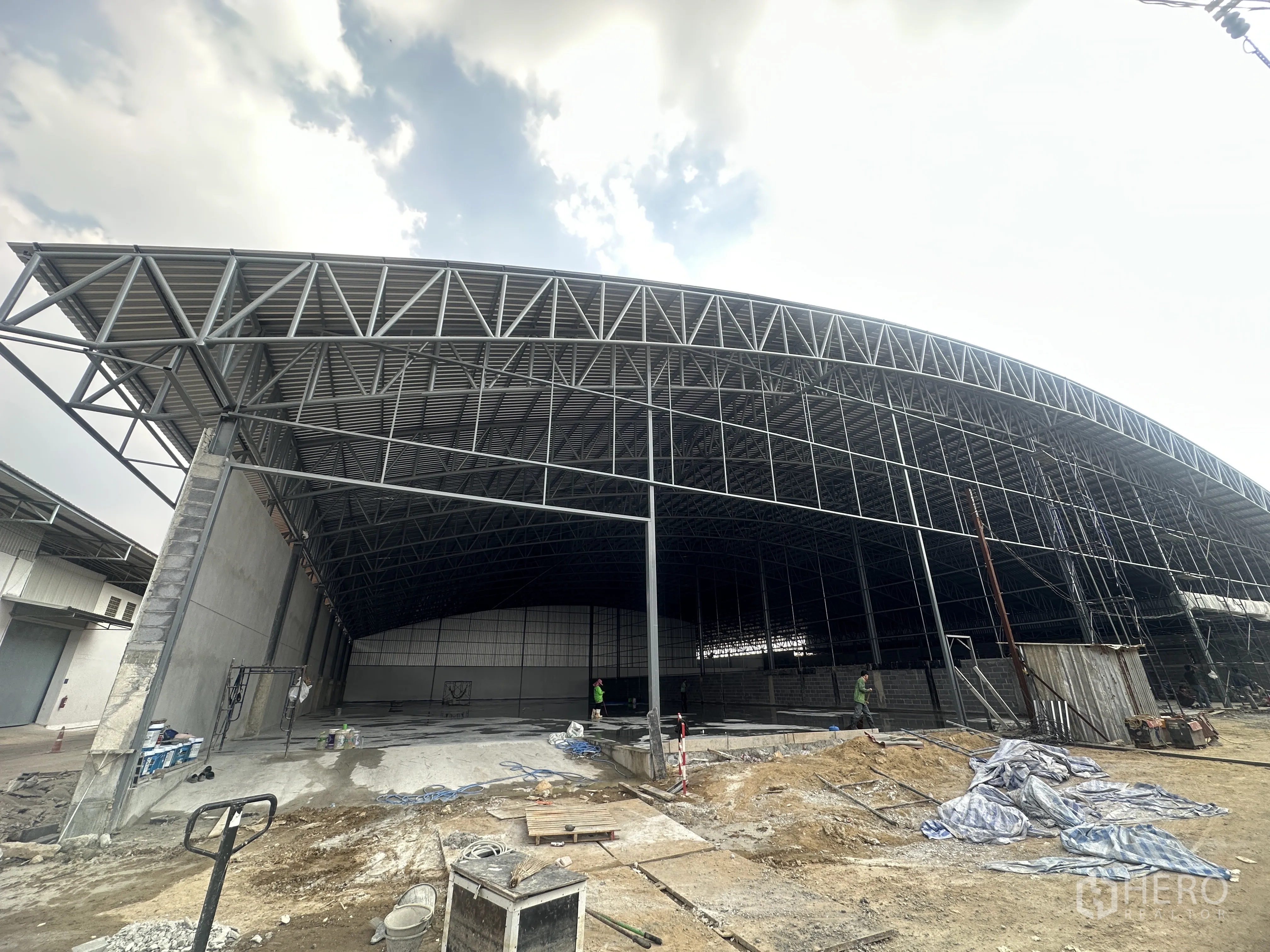 Warehouse for rent in Pak Kret, Nonthaburi - Modern warehouse under construction showing wide-span truss roof and unfinished concrete forecourt.