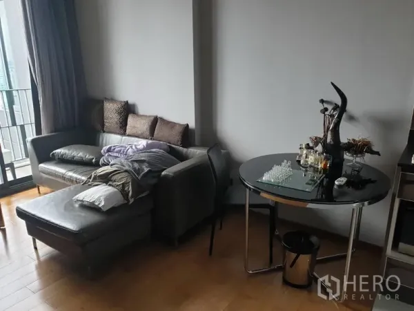 Condominium for rent in Khlong Toei, Bangkok - Cozy corner by the balcony featuring a gray sofa with chaise, round glass-top dining table, and wood flooring.