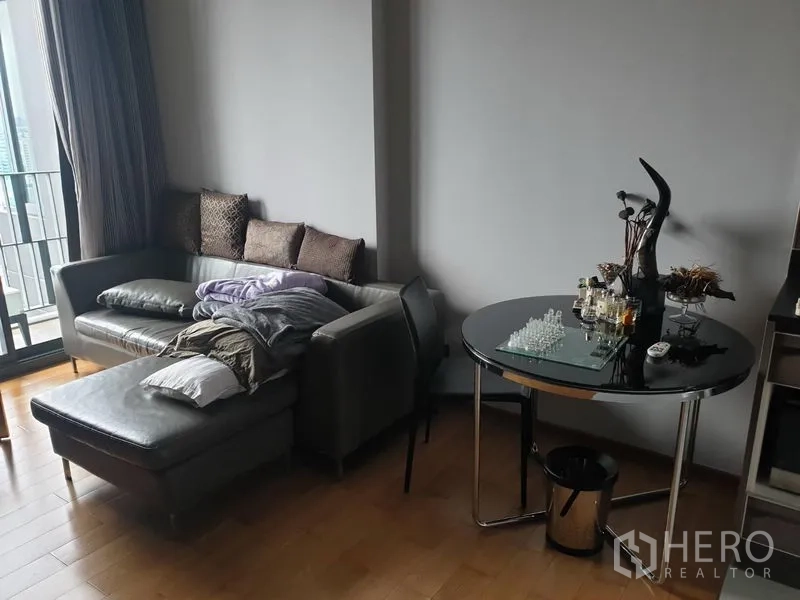Condominium for rent in Khlong Toei, Bangkok - Cozy corner by the balcony featuring a gray sofa with chaise, round glass-top dining table, and wood flooring.