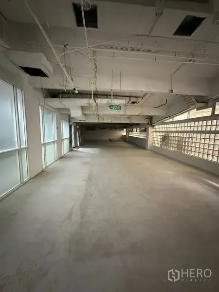 Retail Space for rent - Open-plan retail interior with concrete floor, exposed ceiling ducts, frosted windows and a glass-block wall, with an emergency exit sign.