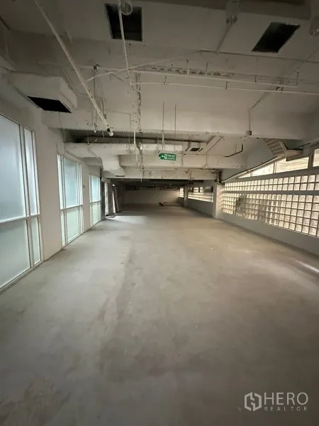 Retail Space for rent - Open-plan retail interior with concrete floor, exposed ceiling ducts, frosted windows and a glass-block wall, with an emergency exit sign.